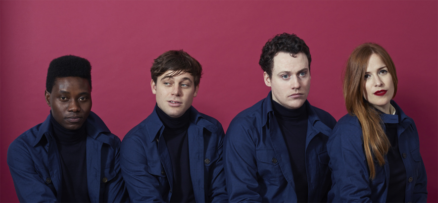 Foto der britischen Band Metronomy – es zeigt drei Männer und eine Frau in blauen Hemden vor einem violetten Hintergund