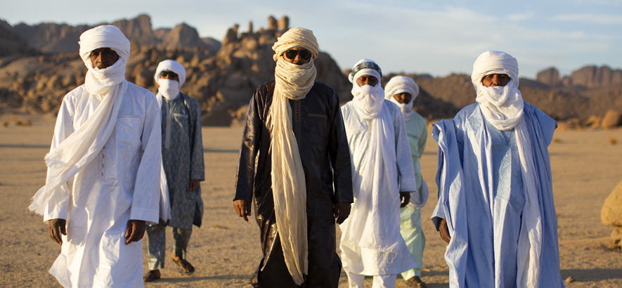 Foto der Tuareg-Band Tinariwen, deren Mitglieder in tradioneller Kleidung nebeneinander durch die Wüste laufen