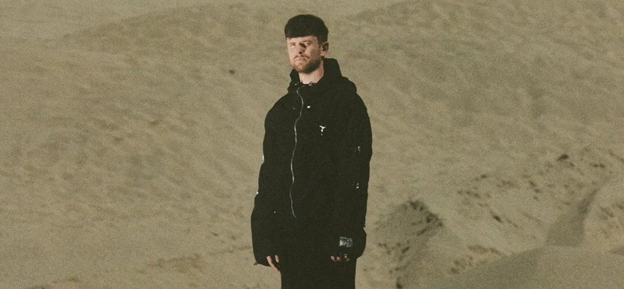 Foto des britischen Musikers James Blake, der allein in schwarzer Kleidung in einer kargen Landschaft steht.