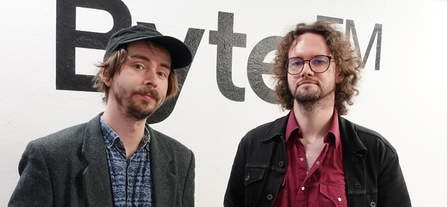 Foto von ByteFM Moderator Marius Magaard und Velvet Bein, Gründer des Musiklabels La Pochette Surprise, die vor einer weißen Wand mit schwarzem ByteFM-Logo stehen